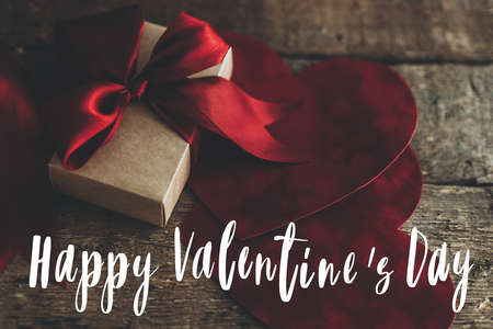Valentines day greeting card. Happy Valentine's day text on stylish gift box with red ribbon and red velvet hearts on rustic wood. Love concept.の写真素材