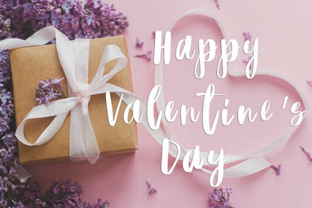 Happy Valentine's day text on lilac flowers, gift box and heart ribbon on pink paper, flat lay. Love concept. Valentines day stylish greeting cardの写真素材