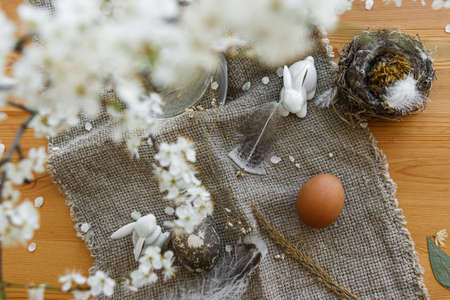 Natural Easter eggs, white bunnies, feather, nest and cherry blooming branch with petals on rustic...