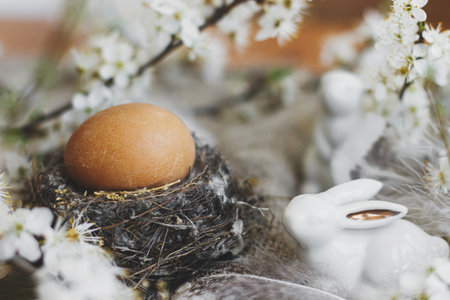 Natural Easter egg in nest and white bunnies, feathers and cherry blooming flowers on rustic linen napkin on table. Happy Easter. Space for text. Eco friendly rural decorの写真素材