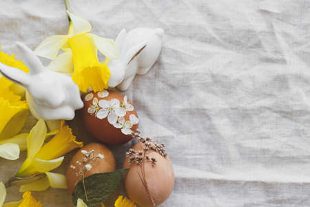 Happy Easter! Stylish Easter eggs decorated with dry flowers and petals on rustic linen napkin with yellow daffodils and white bunnies. Flat lay, Space for text. Creative natural eco friendly decorの写真素材
