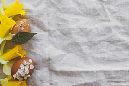 Stylish Easter eggs decorated with dry flowers and petals on rustic linen napkin with yellow daffodils, flat lay. Happy Easter greeting card. Space for text. Creative eco friendly decor of eggsの写真素材