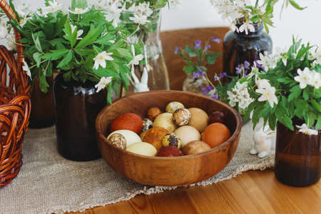 Stylish easter eggs in wooden bowl on rustic table with bunny figurines, wicker basket and spring blooming flowers. Happy Easter! Natural dyed eggs in yellow and red colors.の写真素材