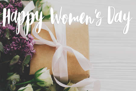 Happy women's day text sign on stylish gift box with lilac and roses flowers on white wood. 8 march International women's day. Stylish floral greeting card, handwritten greetingsの写真素材