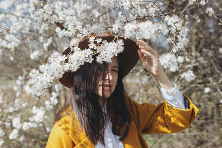 Stylish beautiful woman in hat sensually posing among blooming spring cherry branches outdoor,  calm moment. Portrait of fashionable young brunette female in yellow jacket embracing in white flowersの写真素材