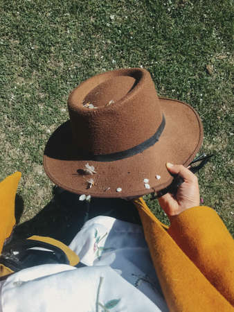 Stylish woman holding hat with white spring petals on background of green grass, top view....