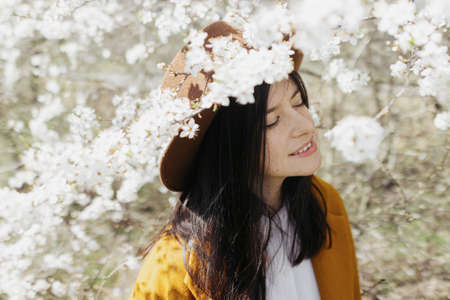 Stylish beautiful woman in hat sensually posing among blooming cherry branches in sunny spring, calm...
