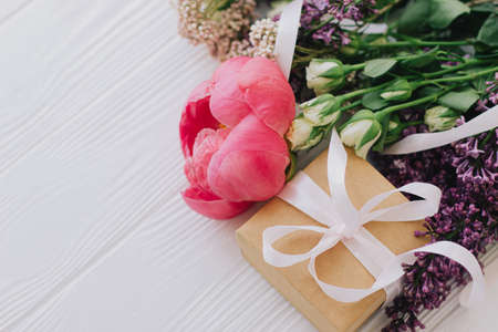 Stylish simple gift box and bouquet with peony, lilac and roses flowers on white wooden background with space for text. Happy mothers day and valentines day concept.の写真素材