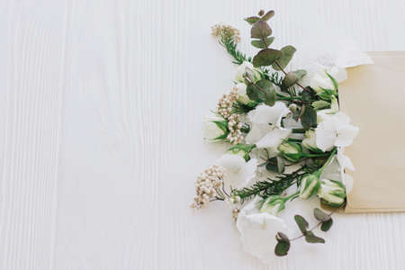 Stylish roses, hydrangea, and eucalyptus branches growing from craft envelope on white wood with...