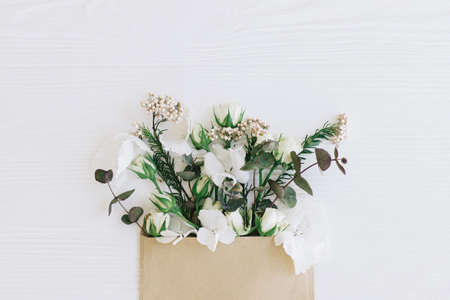 Stylish roses, hydrangea, and eucalyptus branches growing from craft envelope on white wood, flat...