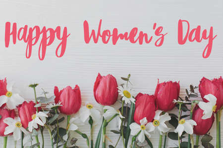 Happy womens day text on beautiful red tulips, daffodils, eucalyptus flowers border on rustic white wood, flat lay. Modern greeting card. Stylish handwritten sign. International women's dayの写真素材