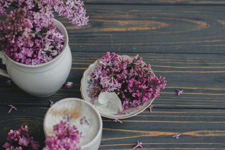 Coffee with lilac petals, blooming lilac branches in vintage cup on dark wooden background. Fresh aromatic coffee in stylish cup and spring flowers on rustic wood. Morning ritualsの写真素材