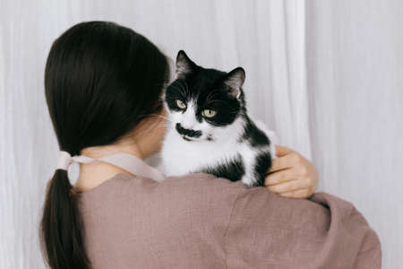 Stylish woman in linen dress holding cute cat on background of pastel fabric. Simple slow living. Young female in boho rustic dress hugging adorable black and white kitty, lovely momentの写真素材