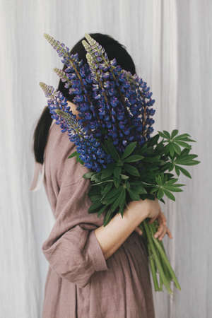 Stylish woman in linen dress holding lupine bouquet on background of pastel fabric. Simple slow living. Young female in boho rustic dress hiding face with lupine wildflowers. Aestheticsの写真素材