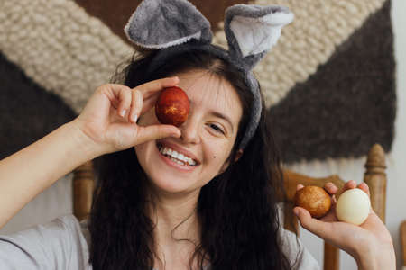 Happy young woman in bunny ears and linen dress holding easter eggs at eyes and smiling on...