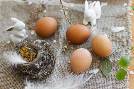 Natural Easter eggs, white bunnies, feathers, nest and cherry blooming branch with petals on rustic linen napkin on table. Happy Easter. Space for text. Eco friendly decorの写真素材