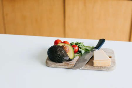 Avocado, cherry tomatoes, arugula, parmesan and knife on wooden cutting board in white modern...