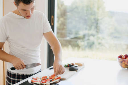 Young man putting tomato slices on bread and making avocado toasts in modern white kitchen. Process of making toasts. Healthy eating and Home cooking conceptの写真素材