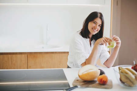 Young happy woman holding green lettuce leaf in hands and smiling in modern white kitchen with fresh fruits and vegetables. Healthy eating and Home cooking concept.の写真素材