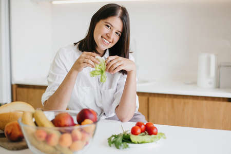 Young happy woman holding green lettuce leaf in hands and smiling in modern white kitchen with fresh fruits and vegetables. Healthy eating and Home cooking concept.の写真素材