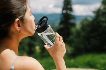 Young woman drinking water from bottle after training on background of sunny mountain hills, face closeup. Outdoor workout. Healthy lifestyle. Sporty female relaxing after training among treesの写真素材
