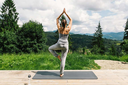 Young fit woman practising yoga and standing in pose on background of sunny mountains hills. Outdoor workout and training. Healthy lifestyle. Sporty Female doing yoga on wooden terrace among treesの写真素材