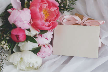 Stylish greeting card, beautiful peonies bouquet, and gift box on soft white fabric on rustic chair. Pink and white peony flowers, present, postcard. Space for text. Happy Mothers dayの写真素材