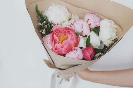 Hand holding beautiful stylish peonies bouquet in paper on background of white wall. Pink and white peony bouquet in female hand. Happy Mothers dayの写真素材