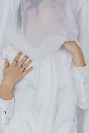 Sensual beautiful woman posing under soft white tulle fabric. Young stylish female in boho floral shirt in modern veil. Mental health. Bridal morningの写真素材