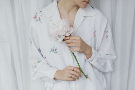 Sensual beautiful woman with pink peony in hands. Young stylish female in boho floral shirt gently holding big peony flower. Tender soft image. Spring aestheticsの写真素材