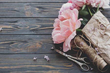 Beautiful stylish peony bouquet in paper, twine and scissors on rustic dark wooden background, copy space. Happy Mothers day greetings. Pink and white peonies flowers and lilac petalsの写真素材