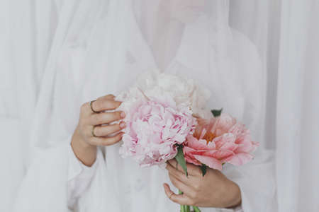 Sensual beautiful woman behind soft white fabric with pink peony in hands. Young stylish female gently holding big pink peony flowers. Tender image. Spring aesthetics. Bridal morningの写真素材