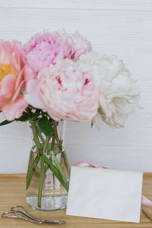 Beautiful peony bouquet in vase, gift box, greeting card and scissors on wooden background, copy space. Happy Mothers day. Pink and white peonies and presentの写真素材