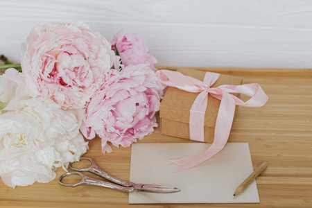 Beautiful peony bouquet, stylish gift box, greeting card and scissors on wooden background. Happy Mothers day. Pink and white peonies and presentの写真素材