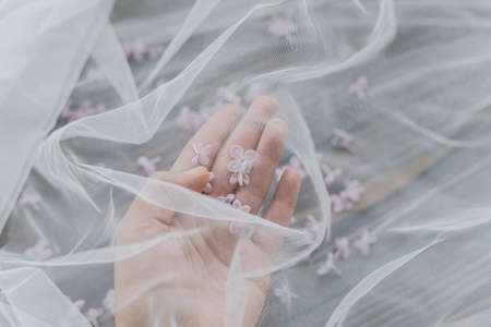 Hand gently holding beautiful lilac petals under soft tulle fabric on dark wooden background, top view with copy space. Tender spring aestheticの写真素材