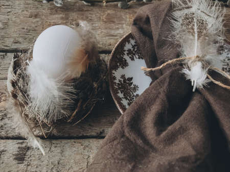 Stylish rustic Easter table setting. Natural easter egg in nest with feathers and napkin on vintage...
