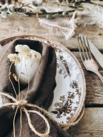 Stylish rustic Easter table setting. Natural easter egg in napkin with flowers, vintage plate and cutlery, soft feathers on aged wooden table. Rural Easter still life. Happy Easterの写真素材
