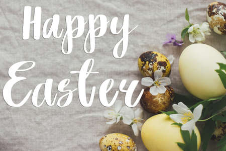 Happy Easter text on modern yellow easter eggs with spring flowers and blossoms on rustic linen background, flat lay, handwritten sign. Beautiful stylish greeting card. Seasons greetingsの写真素材