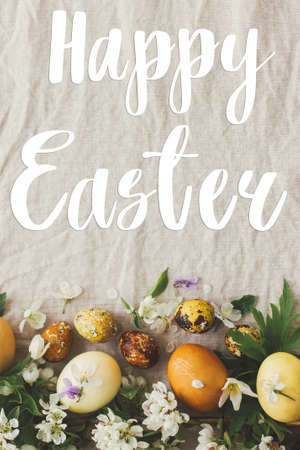 Happy Easter greeting card. Happy Easter text sign handwritten on  modern yellow easter eggs with spring flowers and blossoms on rustic fabric, flat lay. Beautiful postcard, seasons greetingsの写真素材