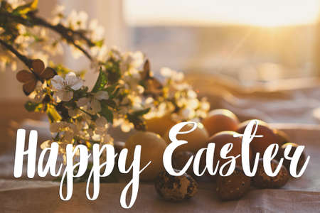 Happy Easter text on modern pastel easter eggs with cherry blossoms on rustic table in sunset light, handwritten sign. Beautiful stylish greeting card. Seasons greetingsの写真素材