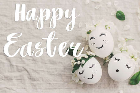 Happy Easter greeting card. Happy Easter text sign handwritten on  eggs with cute funny faces in floral crowns on rustic fabric with spring flowers and blossom. Beautiful postcard, seasons greetingsの写真素材