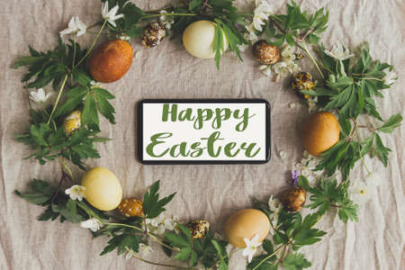 Happy Easter text on phone screen in easter rustic wreath with eggs and flowers, flat lay, handwritten sign. Beautiful stylish greeting card. Seasons greetingsの写真素材