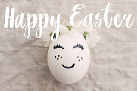 Happy Easter text on egg with cute face in floral crown on rustic fabric with spring flowers, top view, handwritten sign. Beautiful stylish greeting card. Seasons greetingsの写真素材