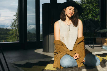 Stylish young woman relaxing in sunny light in modern room. Fashionable hipster female sitting on floor in sunshine on background of window and mountains hillsの写真素材