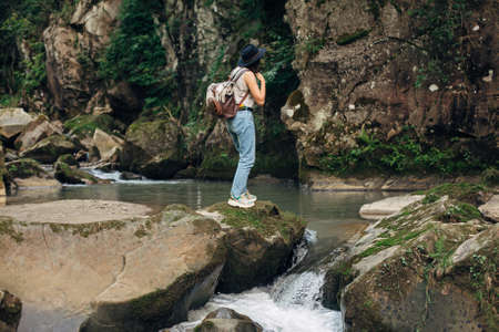 Stylish woman traveler with backpack relaxing on background of river in mountains. Young female in casual cloth and hat standing on rocks at river, back view. Travel and wanderlust. Exploring woodsの写真素材