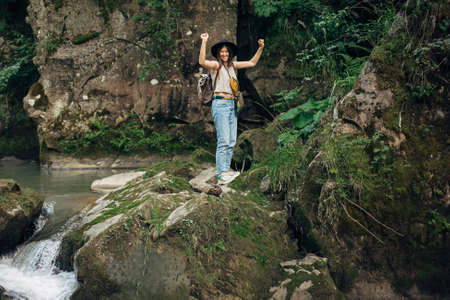 Happy woman traveler with backpack standing on rocks of river in mountains. Young female in casual cloth and hat holding hands up at river, success. Travel and wanderlustの写真素材