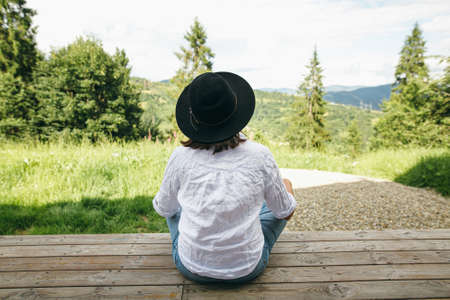 Stylish hipster woman relaxing on wooden terrace on background of sunny mountains hills. Happy young female in hat and casual cloth sitting on porch. Calm tranquil moment. Travel and wanderlustの写真素材