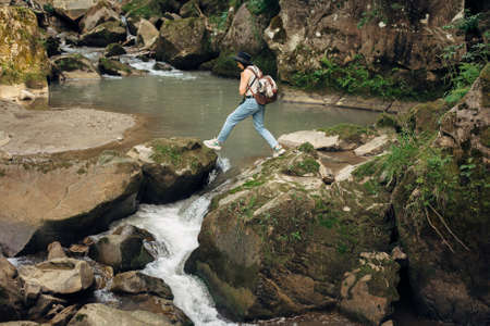 Stylish woman traveler with backpack walking on rocks of river in mountains. Young female in casual cloth and hat jumping across  river. Travel and wanderlust. Hiking and exploring woodsの写真素材