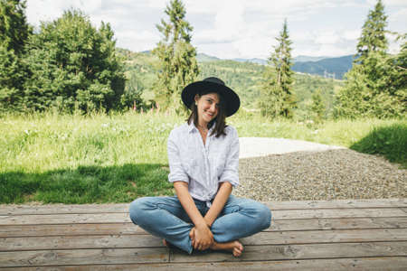 Stylish hipster woman relaxing on wooden terrace on background of sunny mountains hills. Happy young female in hat and casual cloth sitting on porch and smiling. Travel and wanderlustの写真素材