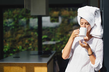 Beautiful young woman enjoying coffee in sunny morning on background of modern kitchen in chalet in mountains. Calm moment. Beautiful female in white pajamas and towel after shower relaxingの写真素材